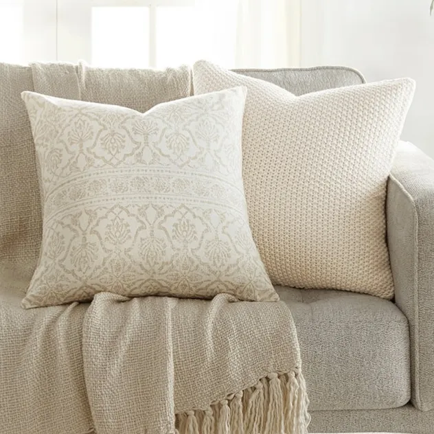 A beige sofa is adorned with two decorative pillows and a fringed throw blanket in neutral tones. A small side table holds a potted plant, adding a touch of greenery to the setting.