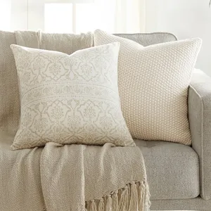 A beige sofa is adorned with two decorative pillows and a fringed throw blanket in neutral tones. A small side table holds a potted plant, adding a touch of greenery to the setting.