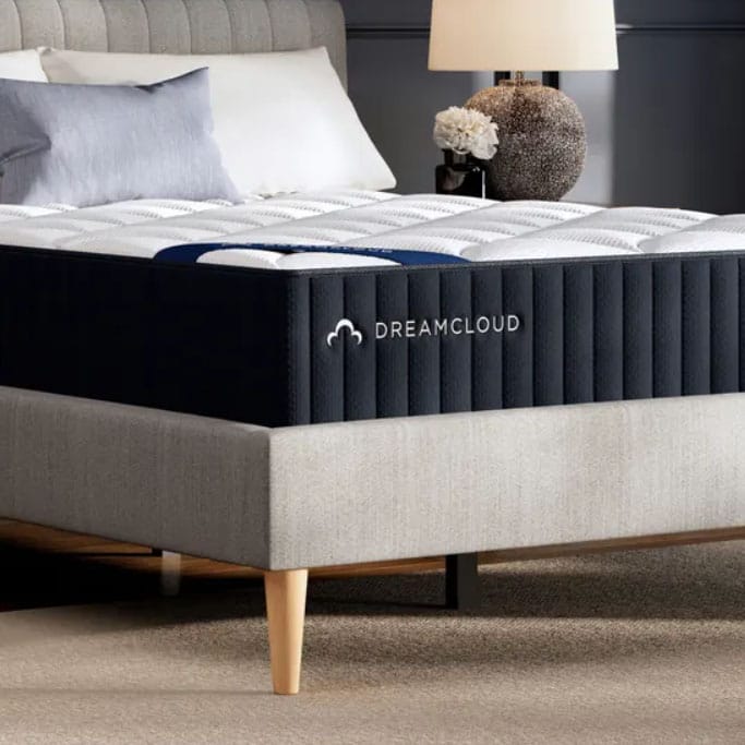 A DreamCloud mattress rests on a modern upholstered bed frame with tapered wooden legs. The setup includes plush bedding and is placed next to a bedside table with a lamp and decorative flowers.