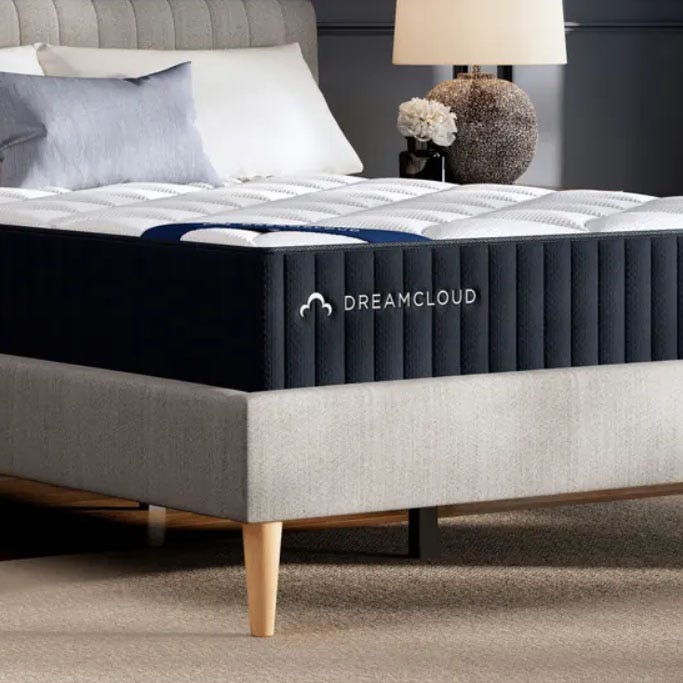A DreamCloud mattress rests on a modern upholstered bed frame with tapered wooden legs. The setup includes plush bedding and is placed next to a bedside table with a lamp and decorative flowers.
