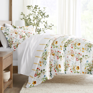 A bed features floral-patterned bedding with matching pillowcases, set against a light and airy background. There is a wooden nightstand with a ceramic item, and a decorative plant adds a natural touch to the room's decor.