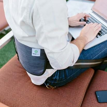 A person sitting with a laptop on their lap, a cushioned lap desk underneath, beside a smartphone on the bench.