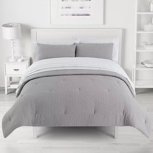 A neatly made bed with a gray quilted comforter, matching pillow shams, and a striped sheet set is placed in a minimalist bedroom featuring a white nightstand with a lamp, and a simple framed artwork above the headboard.