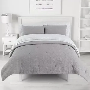 A neatly made bed with a gray quilted comforter, matching pillow shams, and a striped sheet set is placed in a minimalist bedroom featuring a white nightstand with a lamp, and a simple framed artwork above the headboard.