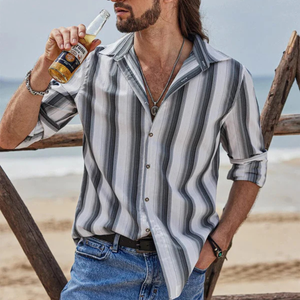 A man on the beach in a striped button-up shirt with rolled sleeves holds a bottle of Corona beer. He's wearing blue jeans, a necklace, and a bracelet.