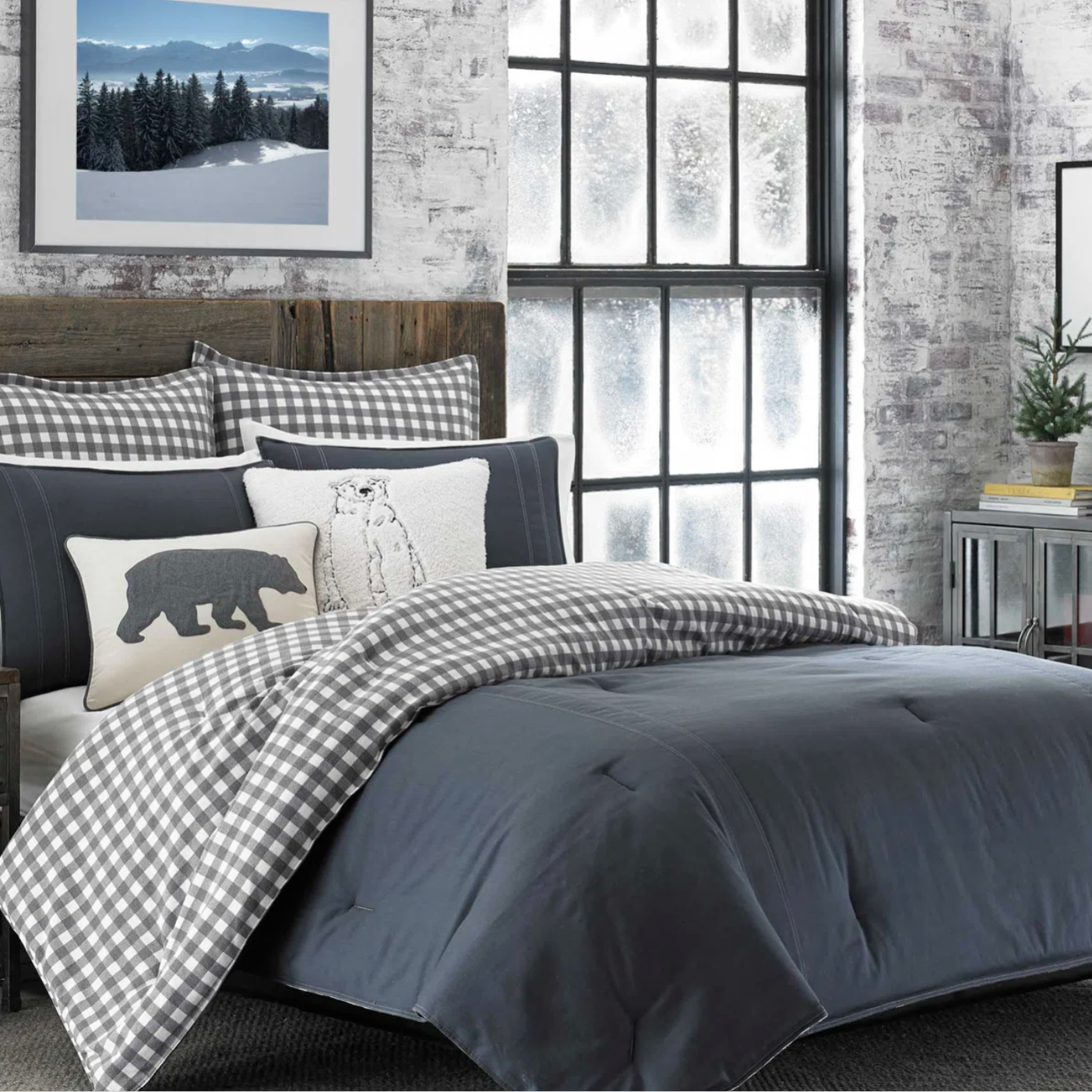 A cozy bedroom setup featuring a dark gray comforter, checkered pillows and duvet, and decorative bear-themed cushions. The wood headboard adds a rustic touch, complemented by a snowy landscape picture on the wall and a frosted window view.