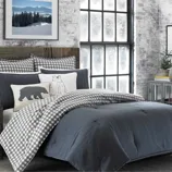 A cozy bedroom setup featuring a dark gray comforter, checkered pillows and duvet, and decorative bear-themed cushions. The wood headboard adds a rustic touch, complemented by a snowy landscape picture on the wall and a frosted window view.