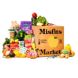 A Misfits Market box is surrounded by a variety of fresh produce, packaged goods like rice and pasta, and canned items.
