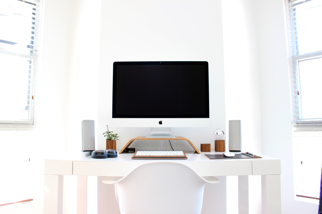 A desktop computer with keyboard and mouse, flanked by two speakers, on a white desk with a monitor stand and small decorative items.