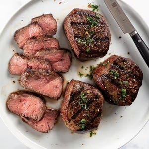 A plate with three grilled steaks garnished with herbs, alongside several slices of medium-rare steak. A knife is placed nearby.