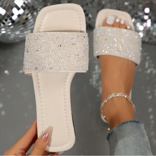 A pair of white slide sandals with sparkling crystal embellishments is shown. One sandal is worn on a foot with painted white toenails and an anklet, while the other is held in a hand displaying manicured nails.
