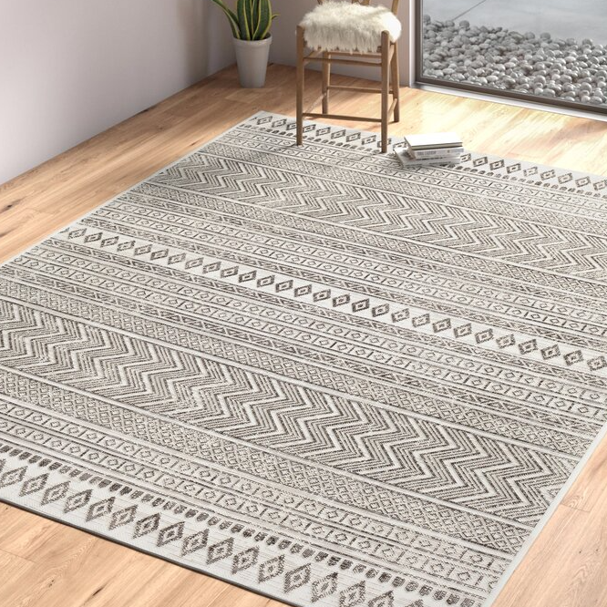 The Laruth Southwestern Rug is 5' x 7', featuring a geometric pattern with zigzags and diamond shapes in neutral tones, set on a wooden floor near a window with a wooden chair and a small plant.