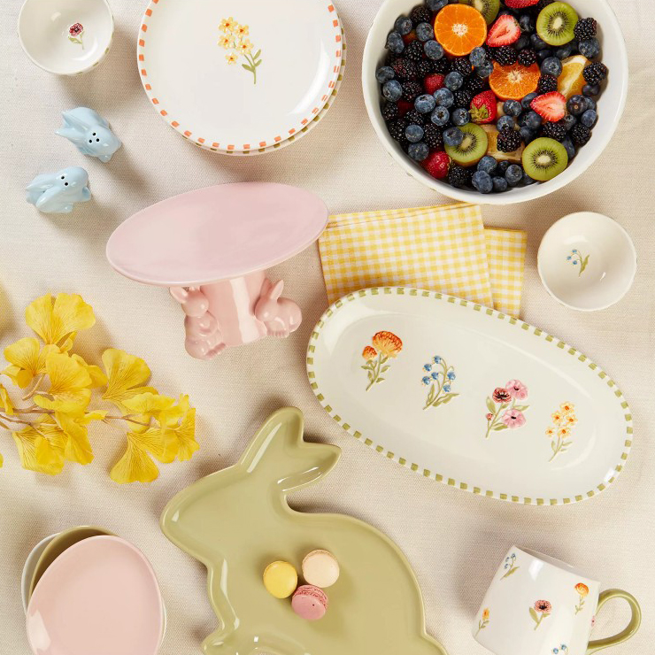 A collection of dishware with floral designs includes a bowl filled with assorted fruits, a yellow-checked napkin, and a green bunny-shaped plate holding macarons, accompanied by yellow flowers and pastel-toned items.