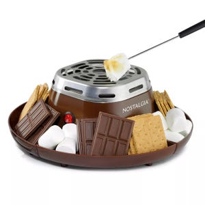 Electric s'mores maker by Nostalgia featuring a roasting station with a skewer, surrounded by marshmallows, chocolate pieces, and graham crackers.
