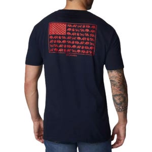 A man wearing a navy T-shirt with a red graphic resembling the American flag, featuring bear silhouettes in place of stripes. The shirt is branded \“Columbia\“ and he has tattoos on his arm.