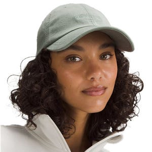 Lululemon Classic Ball Cap features a soft, muted green color with an embroidered logo on the front. The cap has a curved brim and is paired with a white jacket.