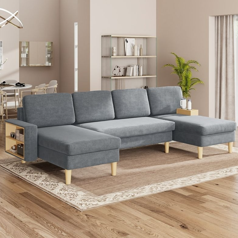 Gray L-shaped sectional sofa with built-in side tables and wooden legs, set in a modern living room with a light rug, hardwood floors, and minimalist decor including a bookshelf and potted plant.