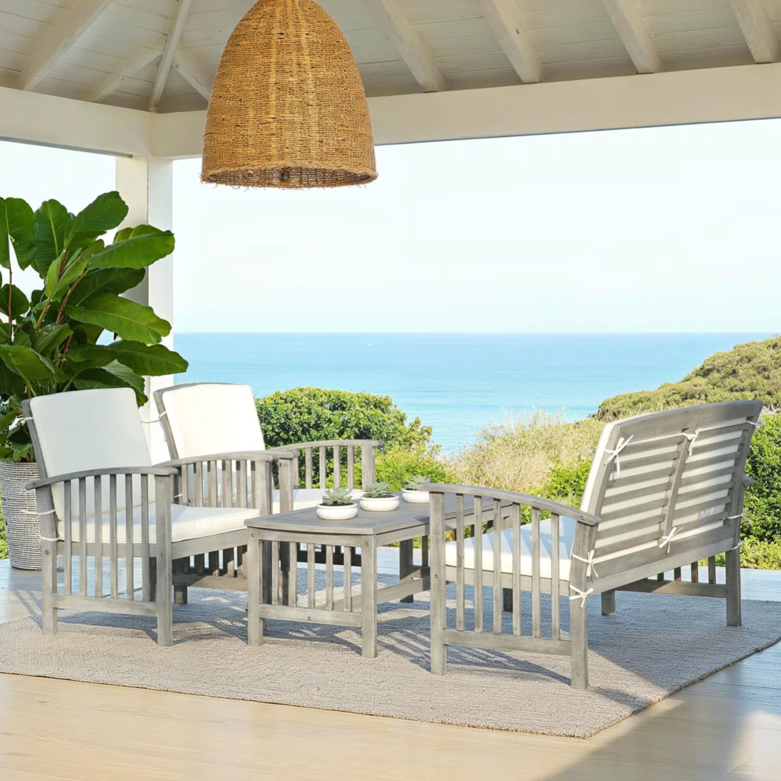 Outdoor patio setup featuring a wooden seating set with white cushions, a coffee table, a woven pendant light, and a large potted plant, set against a backdrop of the ocean and greenery.