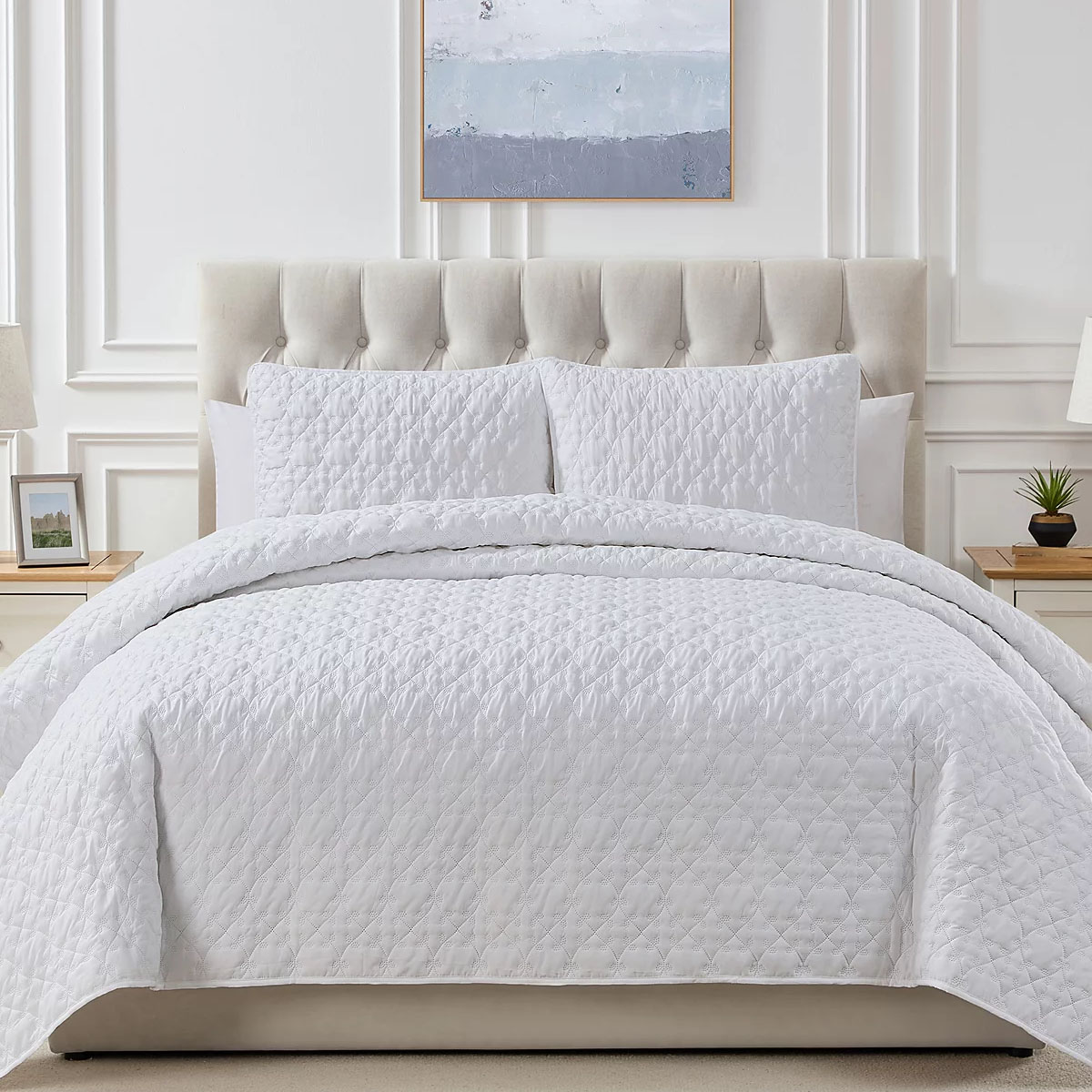 A luxurious quilt set featuring a tufted beige headboard, complete with a white, leaf-stitched quilt and two matching pillow shams, set in an elegant, bright bedroom with minimalist decor, including bedside lamps, a small plant, and a wall painting.