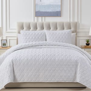 A luxurious quilt set featuring a tufted beige headboard, complete with a white, leaf-stitched quilt and two matching pillow shams, set in an elegant, bright bedroom with minimalist decor, including bedside lamps, a small plant, and a wall painting.