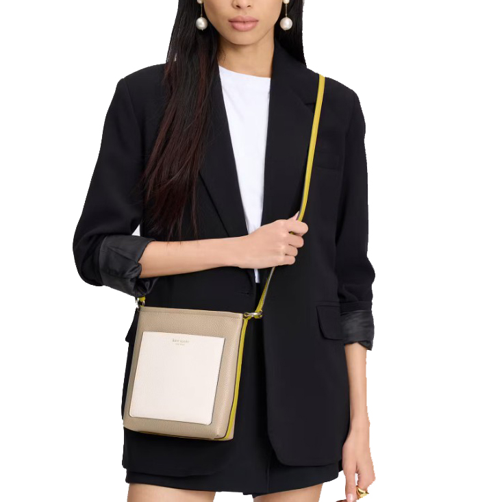A woman is wearing a black blazer and a white T-shirt, carrying a beige and white crossbody bag with a yellow strap. She is also wearing pearl earrings.