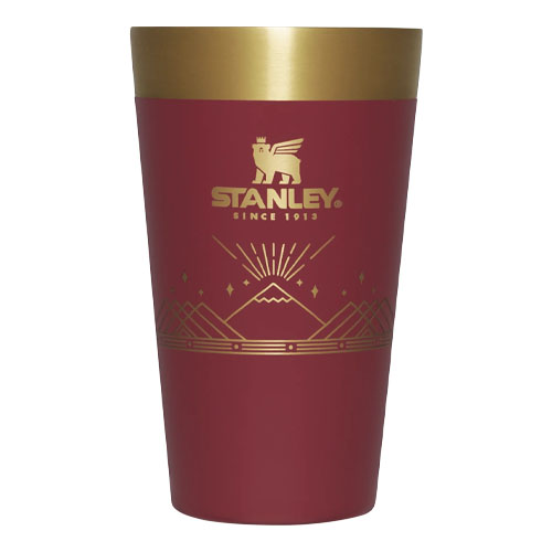 A red Stanley beer pint with a gold rim, featuring the brand's logo and decorative mountain-style graphics.