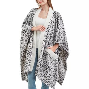 A person is wearing a cozy, leopard-print wearable blanket with a white sherpa lining and large pockets, paired with a white top and jeans.