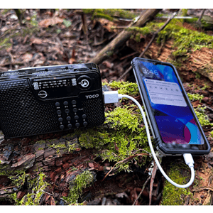 A portable radio and a smartphone connected by a charging cable outdoors on a log.