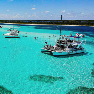 Aerial view of a crystal-clear turquoise beach with two catamarans anchored near a sandbar. People swim and relax in the water, surrounded by flags and snorkeling gear.