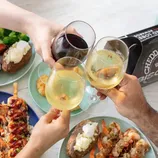 Three people toast with glasses of white and red wine over plates of lobster tails, shrimp, baked potatoes, broccoli, and barbecue items, accompanied by Cheddar Bay Biscuits.