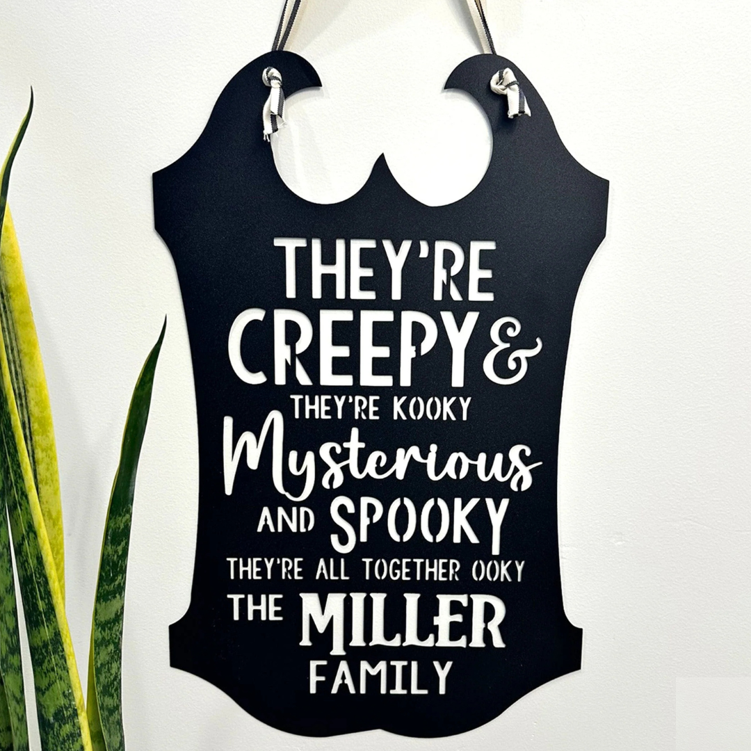 A black hanging plaque features the phrase \“They're creepy & they're kooky, mysterious and spooky, they're all together ooky, the *Miller* family,\“ inspired by a famous theme song.
