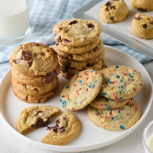 Assorted cookies on a plate, featuring chocolate chunk and colorful sprinkles varieties. A glass of milk and a baking sheet with more cookies are in the background.