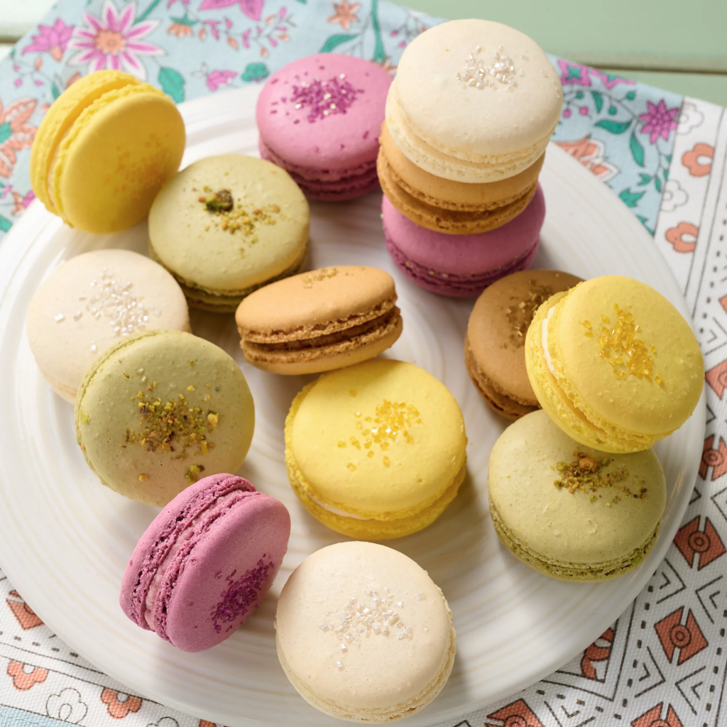 A plate of colorful macarons in shades of yellow, green, pink, and beige is arranged on a floral-patterned cloth. Each macaron is topped with decorative sprinkles or nuts.