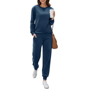 A woman wears a navy blue 2-piece loungewear set, featuring a long-sleeve top and jogger-style pants with pockets. She carries a brown shoulder bag, holds a coffee cup, and wears white sneakers.