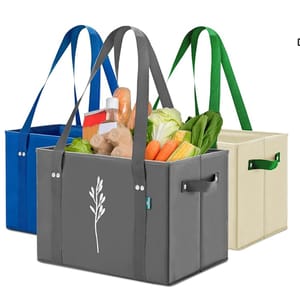 Three reusable tote bags in blue, gray, and beige colors with sturdy handles and side handles. The gray bag contains groceries like lettuce, carrots, and corn.