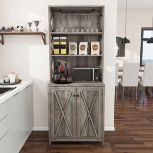 Farmhouse-style kitchen pantry with wood finish, featuring glassware on top, four boxes of pasta or grains on the second shelf, a microwave and assorted bottles on the third, and storage cabinets below. A decorative wall shelf with trophies and decorations is mounted nearby.