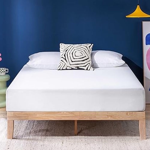 A bed with a wooden frame features a white mattress topped with two white pillows and a black-and-white geometric patterned cushion. A blue wall, colorful artwork, and a yellow pendant light complete the modern room decor.