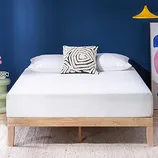 A bed with a wooden frame features a white mattress topped with two white pillows and a black-and-white geometric patterned cushion. A blue wall, colorful artwork, and a yellow pendant light complete the modern room decor.