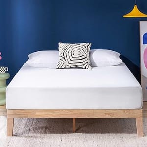 A bed with a wooden frame features a white mattress topped with two white pillows and a black-and-white geometric patterned cushion. A blue wall, colorful artwork, and a yellow pendant light complete the modern room decor.