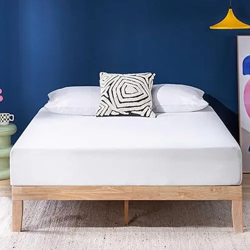 A bed with a wooden frame features a white mattress topped with two white pillows and a black-and-white geometric patterned cushion. A blue wall, colorful artwork, and a yellow pendant light complete the modern room decor.