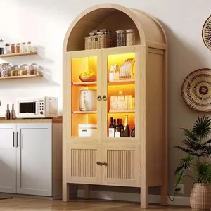 A wooden cabinet with glass doors is illuminated, displaying dishes, glasses, and bottles. Shelves above hold jars, and a countertop features a microwave and decor.