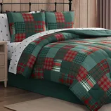 A bed features a patchwork quilt and matching pillow shams with red and green plaid patterns, complemented by white sheets with small black tree designs. The setup includes a black metal headboard and a wooden nightstand.