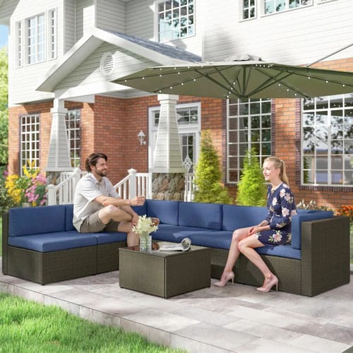 Outdoor patio set with blue cushioned sectional seating and a wicker coffee table, underneath a large green umbrella, situated on a stone patio in front of a house.