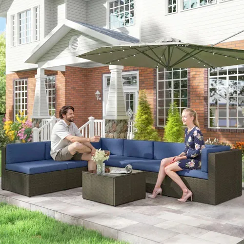 Outdoor patio set with blue cushioned sectional seating and a wicker coffee table, underneath a large green umbrella, situated on a stone patio in front of a house.