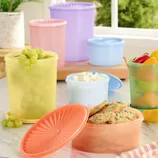 A variety of colorful, round plastic storage containers are shown, each filled with items like grapes, marshmallows, cookies, and sliced vegetables, with lids either on or beside them.