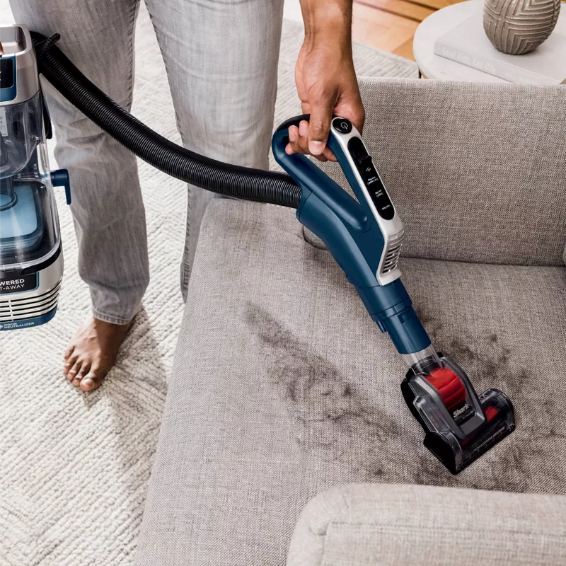 A person is using a handheld upholstery vacuum cleaner to clean a grey sofa.