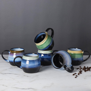 Set of six ceramic mugs with blue, green, and beige gradient coloring, displayed on a marble surface. Coffee beans are scattered nearby.