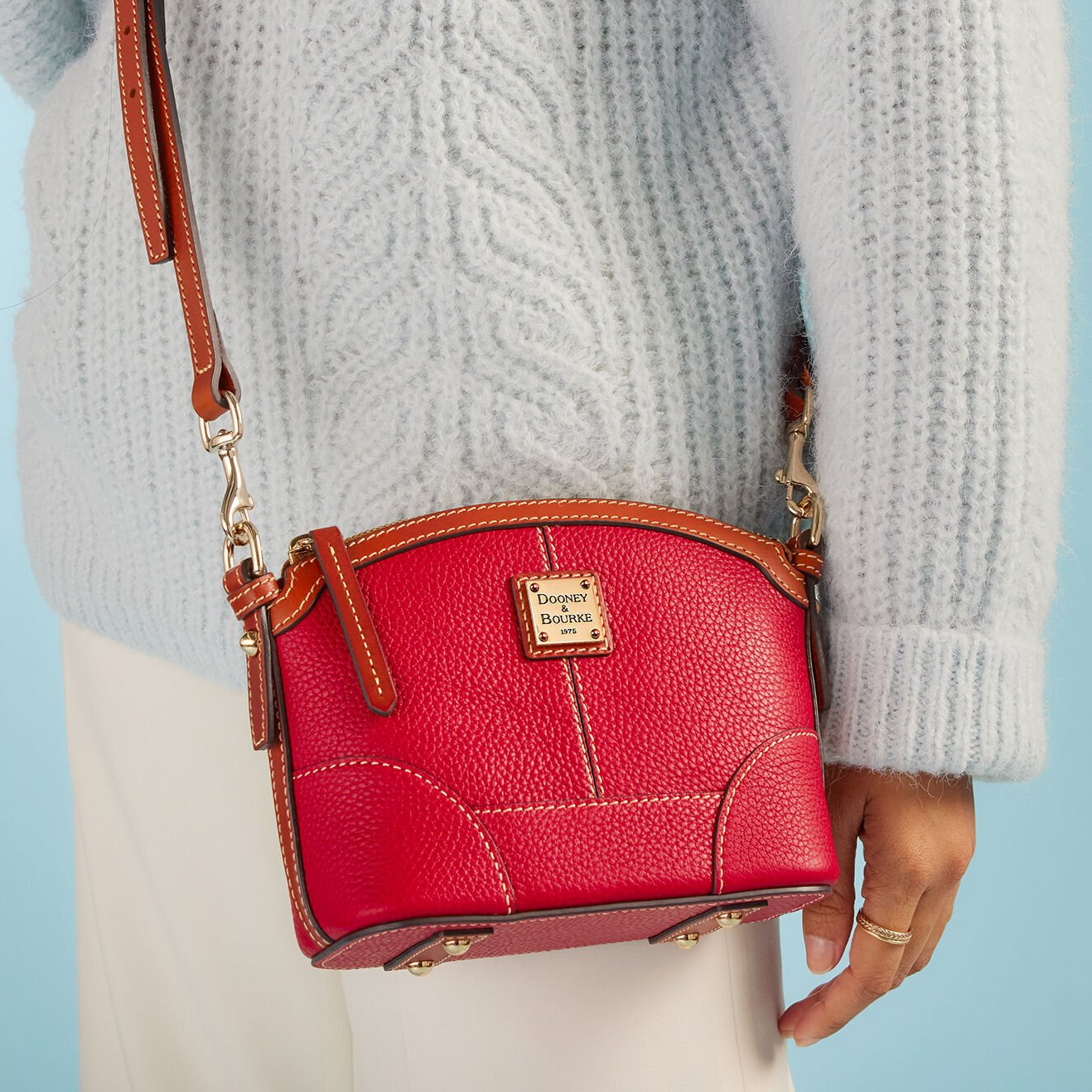 A red Dooney & Bourke handbag with brown trim and a detachable strap, held by a person wearing a light blue sweater.