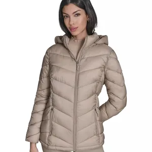 A woman is wearing a beige hooded puffer jacket with a front zipper and two zippered pockets. The jacket features a chevron quilted pattern.