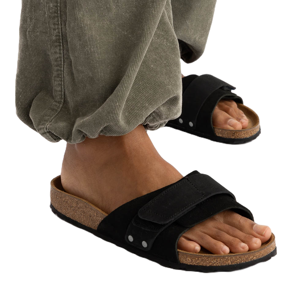 A person wearing green corduroy pants and black open-toe sandals with a cork sole and adjustable strap.